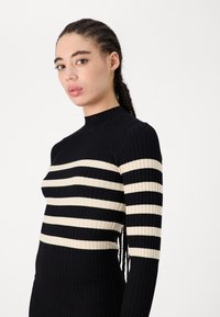 Pulover iz ribane pletenine s črnimi in kremnimi vodoravnimi črtami, visokim ovratnikom in prilegajočim se dizajnom. Teksturirana tkanina z dolgimi rokavi.
