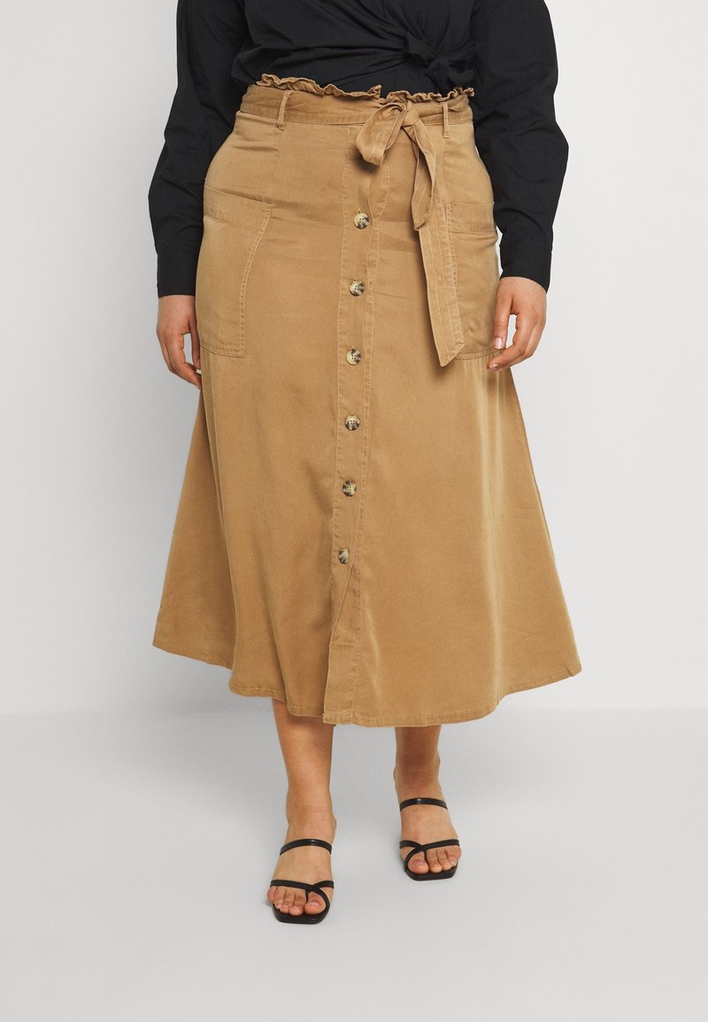 Femme portant une jupe midi beige boutonnée devant avec des poches avant et une ceinture nouée à la taille, associée à un top noir à manches longues et des sandales noires.