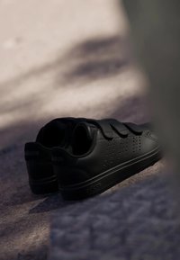 Chaussures de sport noires avec une texture en cuir lisse, trois bandes Velcro, des détails perforés et une semelle en caoutchouc plate.