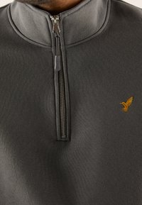 Jersey gris con diseño de media cremallera, que presenta una tela texturizada. Un logo bordado de un pájaro dorado está ubicado en el pecho izquierdo.