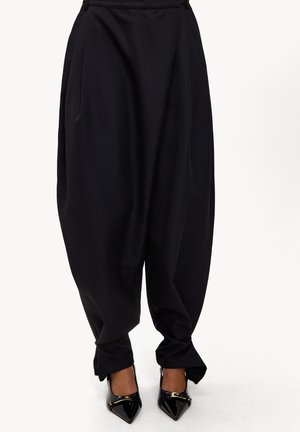 Pantalons noirs amples avec entrejambe abaissé et chevilles fuselées associés à des escarpins noirs en vernis avec des accents dorés.