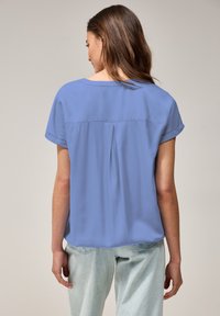 Blouse bleue à manches courtes avec un détail de pli au dos, fabriquée en tissu léger, associée à un jean délavé, offrant une coupe décontractée.