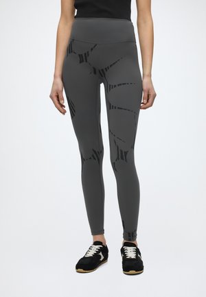 Γυναίκα που φορά γκρι ψηλόμεσα κολάν με μαύρα αφηρημένα σχέδια και μαύρα αθλητικά παπούτσια με λευκά κορδόνια, στέκεται μπροστά σε λευκό φόντο.