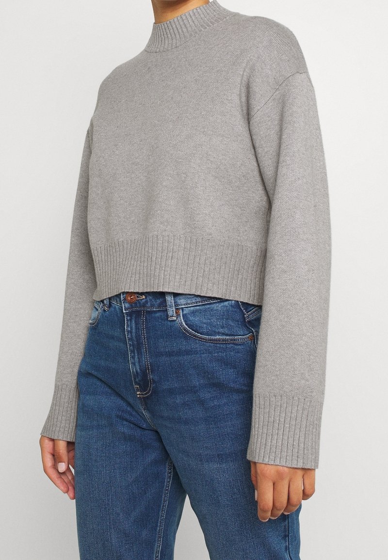 Personne portant un pull court en maille côtelée gris clair à manches longues et un jean bleu taille haute sur un fond uni.