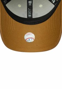 Bronzefarbener Hut mit schwarzem Schirm, der auf der Vorderseite ein rundes Baseball-Abzeichen trägt. Strukturiertes Gewebe mit sichtbaren Nähdetails.