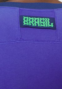 Fialový sportovní dres s texturovanou tkaninou. Obsahuje modrou nášivku na výstřihu s nápisem "BRASIL" vytištěným zeleným stylizovaným písmem.
