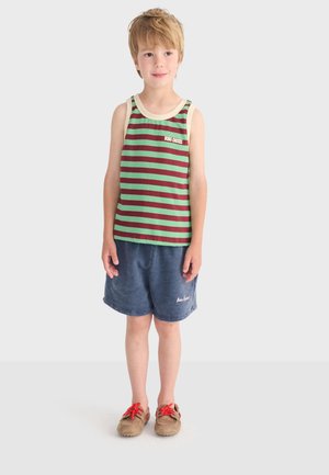 Garçon blond debout, portant un débardeur rayé vert et rouge, un short bleu et des chaussures marron à lacets rouges, sur un fond gris clair.