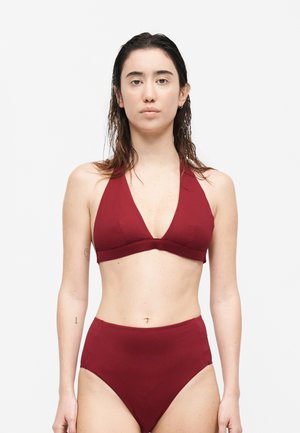 Γυναίκα με βρεγμένα μαλλιά που φοράει μαγιό δύο τεμαχίων μπορντό, στέκεται μπροστά σε απλό λευκό φόντο, αντικρίζοντας μπροστά.