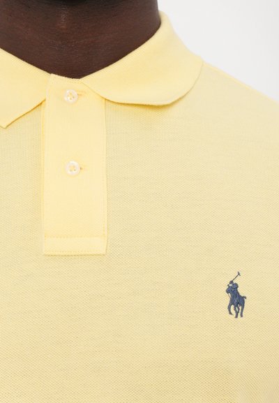 Dzeltena polo krekla izgatavots no teksturēta auduma, ar apkakli, diviem pogām un mazu tumši zilu logotipu ar jātnieku kreisajā krūtīs.