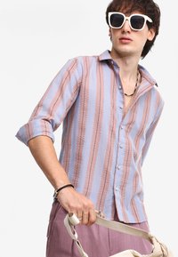 Camisa de botones con rayas verticales alternas en rosa y azul claro, con una tela texturizada y mangas enrolladas. Combinada con un bolso claro.