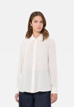 Donna che indossa una camicetta bianca trasparente a maniche lunghe con collo e bottoni, abbinata a jeans blu scuro, in piedi davanti a uno sfondo semplice.