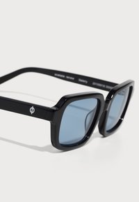 Ochelari de soare din plastic negru cu lentile albastre, cadru pătrat și detalii subtile ale logo-ului pe brațele ochelarilor. Textură netedă și finisaj lucios.