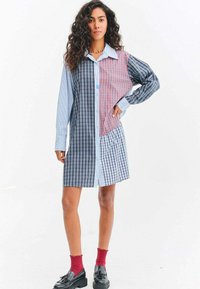 Mladá žena v barevných kostkovaných šatech ze záplat, červených ponožkách a černých mokasínách, stojící s jednou rukou v bok na bílém pozadí.