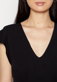 Nainen, joka pukeutuu mustaan V-kaula-aukkoiseen paitaan lyhyillä hihoilla, paljastaen kaulan, solisluun ja ylärinnan valkoista taustaa vasten.