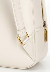 Borsa color crema realizzata in materiale sintetico liscio. Presenta una cerniera dorata, un accento con logo e dettagli cuciti sulla tracolla.