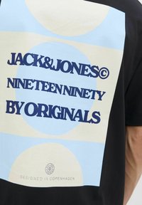 Sort T-shirt med et lysblåt grafisk design med cirkulære former og fed tekst: "JACK&JONES NINETEEN NINETY BY ORIGINALS" på ryggen.