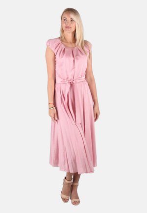 Blondes Frau, die ein hellrosa ärmelloses Midikleid mit Taillenschnürung und beige Knöchelriemenabsätze trägt, steht vor einem weißen Hintergrund.