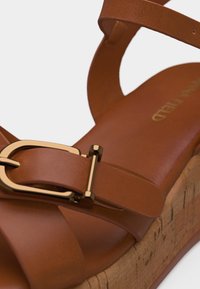 Καφέ δερμάτινο πέδιλο πλατφόρμα με φελλό στη σόλα και αγκράφα σε χρυσό τόνο στο λουράκι του αστραγάλου, σε κοντινή λεπτομέρεια.