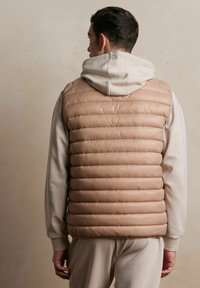 Beżowa, pikowana kamizelka z poziomymi szwami, noszona na jasnoszarym hoodie. Gładka faktura, lekka konstrukcja i praktyczne warstwowe noszenie.