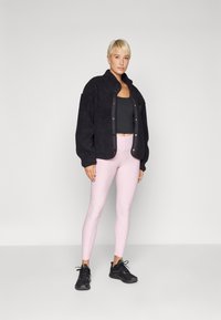 Sort fleecejakke med lommer, parret med en sort crop top og lyserøde leggings. Model iført sorte træningssko.