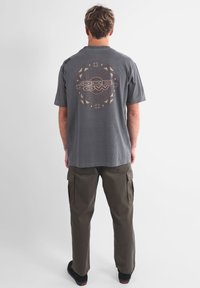 T-shirt en coton gris à manches courtes, avec un motif graphique circulaire dans le dos. Associé à un pantalon cargo olive et des chaussures noires.