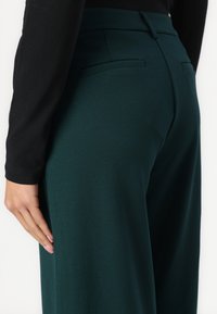 Dark green trousers with a smooth texture, featuring two back pockets and a tailored fit. The waistband is flat and structured.