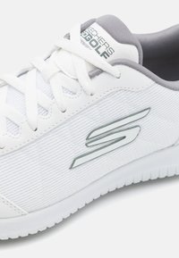 Vit sneaker med en texturerad ovandel, grå detaljer och en reflekterande logotyp. Har vita snörningar och en gummisula för bra grepp.