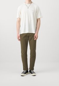 Vit t-shirt med kort ärm och subtilt tryck, kombinerad med olivgröna slim-fit byxor och svarta sneakers med vita sulor.