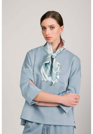 Joven con cabello oscuro recogido, vestida con un top azul claro y pantalones a juego, con un pañuelo estampado atado alrededor del cuello.