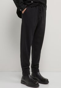 Person, der schwarze Jogginghosen und schwarze Lederstiefeletten trägt, steht vor einem schlichten hellen Hintergrund.