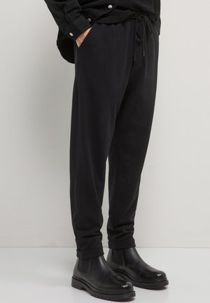 Person, der schwarze Jogginghosen und schwarze Lederstiefeletten trägt, steht vor einem schlichten hellen Hintergrund.