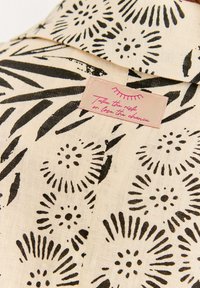 Světle béžová tkanina s černým květinovým vzorem. Obsahuje štítek s růžovým textem, který říká: "Vezmi si riziko nebo ztrácej šanci."