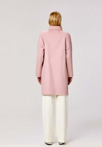 Manteau en laine rose clair avec un col, silhouette droite et manches longues, associé à un pantalon large blanc. Texture lisse, peu de détails.