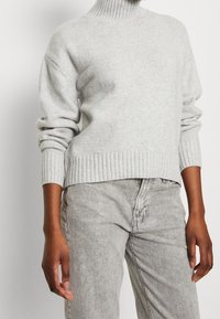Pull à col roulé surdimensionné gris clair en maille douce avec poignets et ourlet côtelés. Associé à un jean gris délavé, présentant une coupe décontractée.