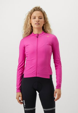 Frau mit lockigem Haar, die eine enganliegende, langärmlige, leuchtend pinke Radjacke und schwarze gepolsterte Radhosen mit reflektierenden Streifen trägt.