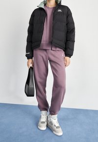 Svart vadderad jacka med en ljusgrön innervikt, i kombination med en lavendeltröja och matchande sweatshirtbyxor. Vita sneakers och svart handväska.