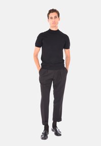 Giovane uomo che indossa un maglione a collo alto nero a maniche corte, pantaloni neri su misura, calzini neri e mocassini neri, in piedi con le mani in tasca.