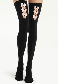 Jambieres noires montantes avec cinq nœuds roses le long du côté externe, offrant une texture douce et transparente ainsi qu'une finition lisse.