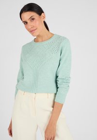 Maglione lavorato a maglia di colore verde chiaro con un motivo a rombi testurizzato, scollo rotondo e polsini a costine, abbinato a pantaloni color crema.