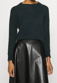Tmavě zelený pletený svetr s kulatým výstřihem a dlouhými rukávy, kombinovaný s černou koženou sukní. Svetr má volný střih a texturovaný povrch.