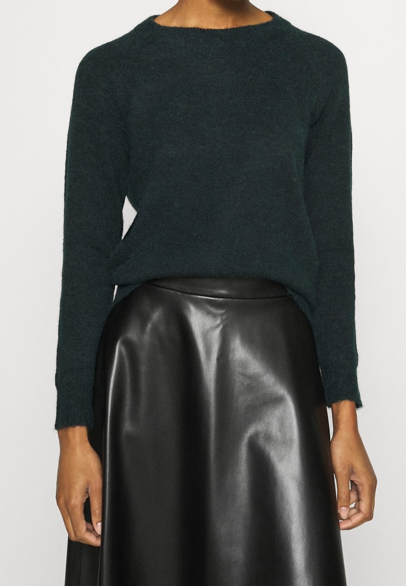 Tmavě zelený pletený svetr s kulatým výstřihem a dlouhými rukávy, kombinovaný s černou koženou sukní. Svetr má volný střih a texturovaný povrch.