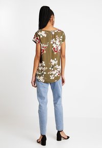 Oliven grøn blomstret bluse med korte ærmer, præget af hvide og røde blomstermønstre. Den har en rundt nederste kant og en nøglehul detalje bagpå.