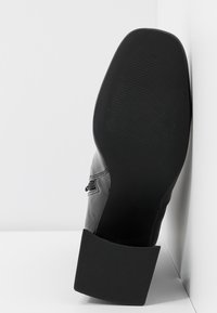 Chaussure noire à finition brillante, dotée d'une semelle en caoutchouc texturée et d'un talon carré. Le côté présente un détail de fermeture éclair.