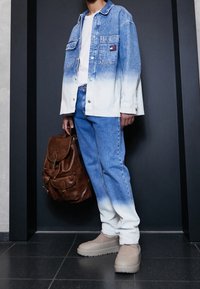 Osoba v modro-bílém ombre džínsovém saka a džínách, v béžových mokasínech, držící hnědou koženou batoh, stojící proti tmavé zdi.