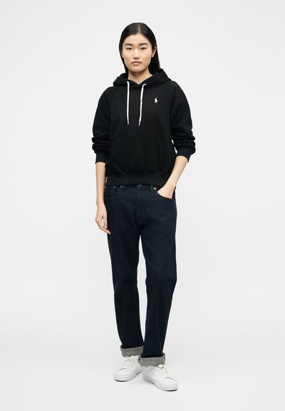 Νεαρή γυναίκα που στέκεται φορώντας μαύρη μπλούζα με κουκούλα και λευκό λογότυπο, σκουρόχρωμο τζιν με γυρισμένα μανίκια και λευκά αθλητικά παπούτσια, τα χέρια χαλαρά, ουδέτερο φόντο.