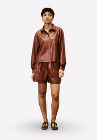 Femme de pieds à tête portant une chemise en simili cuir marron à boutons et un short assorti, avec des chaussures T-strap marron, sur un fond blanc.