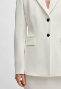 White blazer featuring a tailored fit, sharp lapels, and two black buttons, with front pockets and a smooth textured fabric.