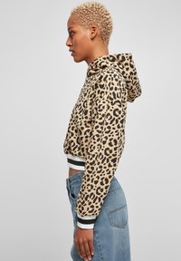 Beige avkuttet hettegenser med svart leopardmønster. Har hette, elastiske mansjetter og en hvit-svart stripete søm. Fiberaktig tekstur.