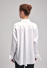 Camicia bianca in cotone con collo a punta, vestibilità ampia, fessure laterali e dettagli con fiocco ai polsini. Texture liscia, design semplice e senza motivi.