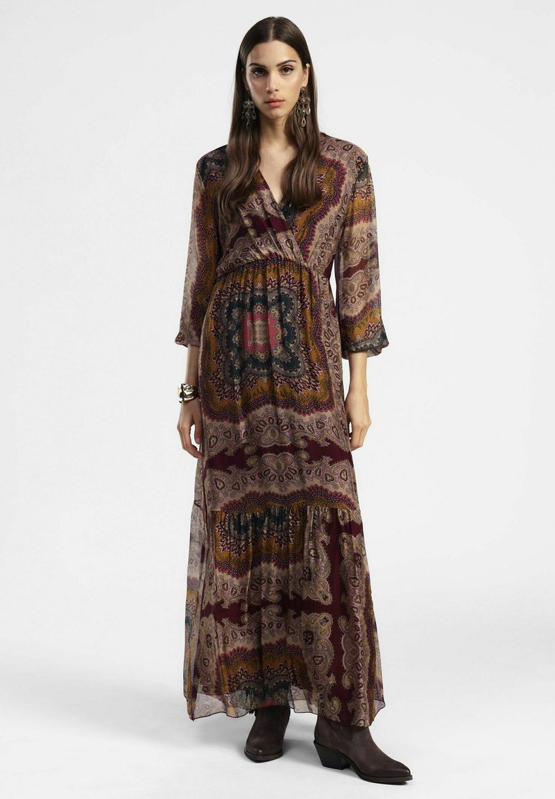 Abito lungo stampato con vari motivi paisley in bordeaux, marrone e blu. Forma fluida con maniche lunghe e scollatura a V, abbinato a stivali marroni.
